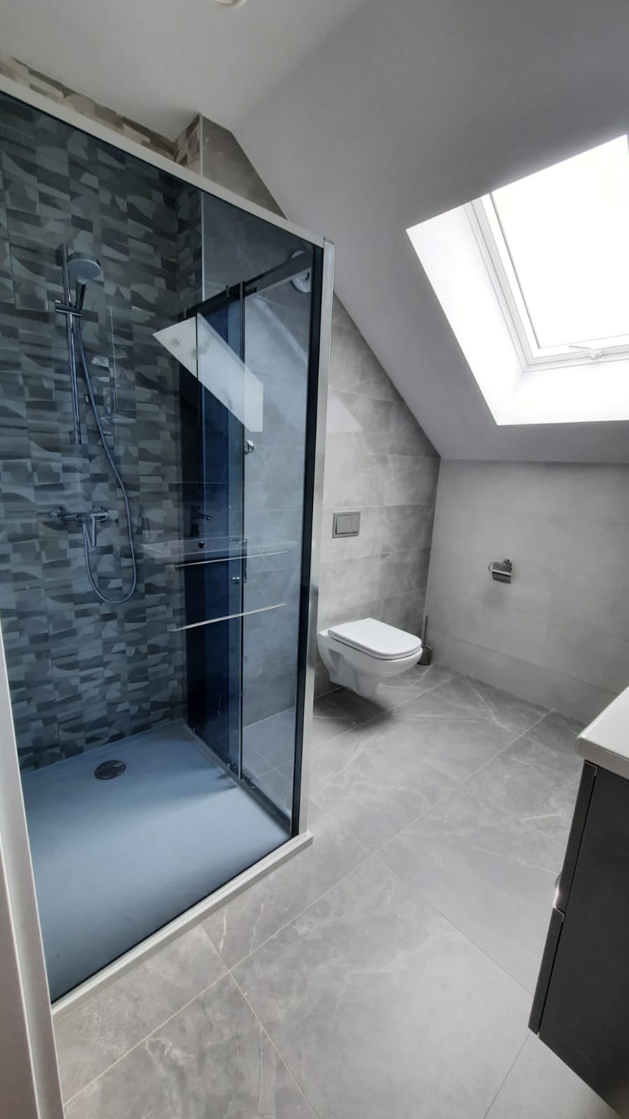 Nowoczesna łazienka na poddaszu z kabiną prysznicową typu walk-in, wiszącą toaletą i szarą glazurą imitującą marmur, doświetlona oknem dachowym.