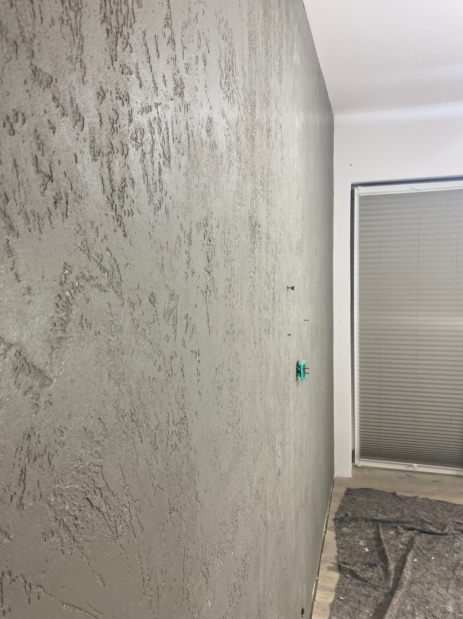 Szara ściana z dekoracyjną strukturą, widoczne gniazdko elektryczne w trakcie montażu i fragment okna z żaluzjami.