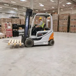 Operator w wózku widłowym STILL RX 60-25, w żółtym kasku i kamizelce odblaskowej, przewozi pustą paletę w magazynie z zapakowanymi kartonami na paletach.