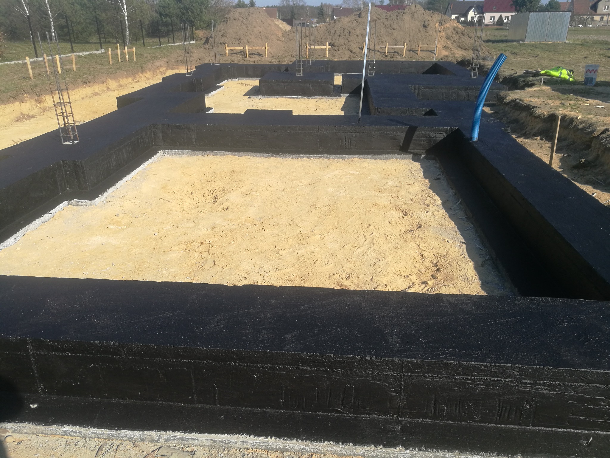 Zabezpieczone hydroizolacją fundamenty domu jednorodzinnego, widoczne zbrojenia i niebieska rura instalacyjna, piasek wewnątrz wykopu.