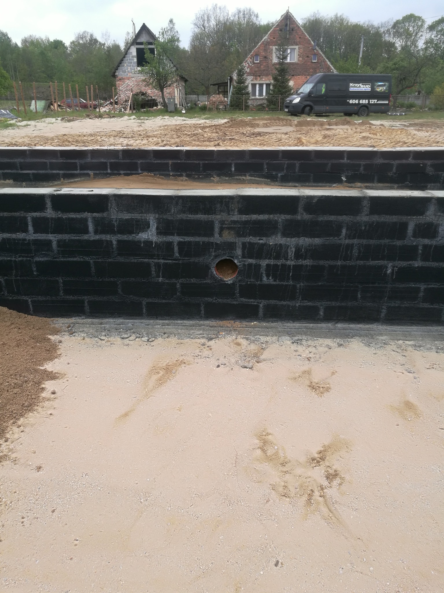Czarne bloczki fundamentowe z otworem na rurę, widoczne piasek, częściowo zrujnowany budynek i firmowy van w tle.