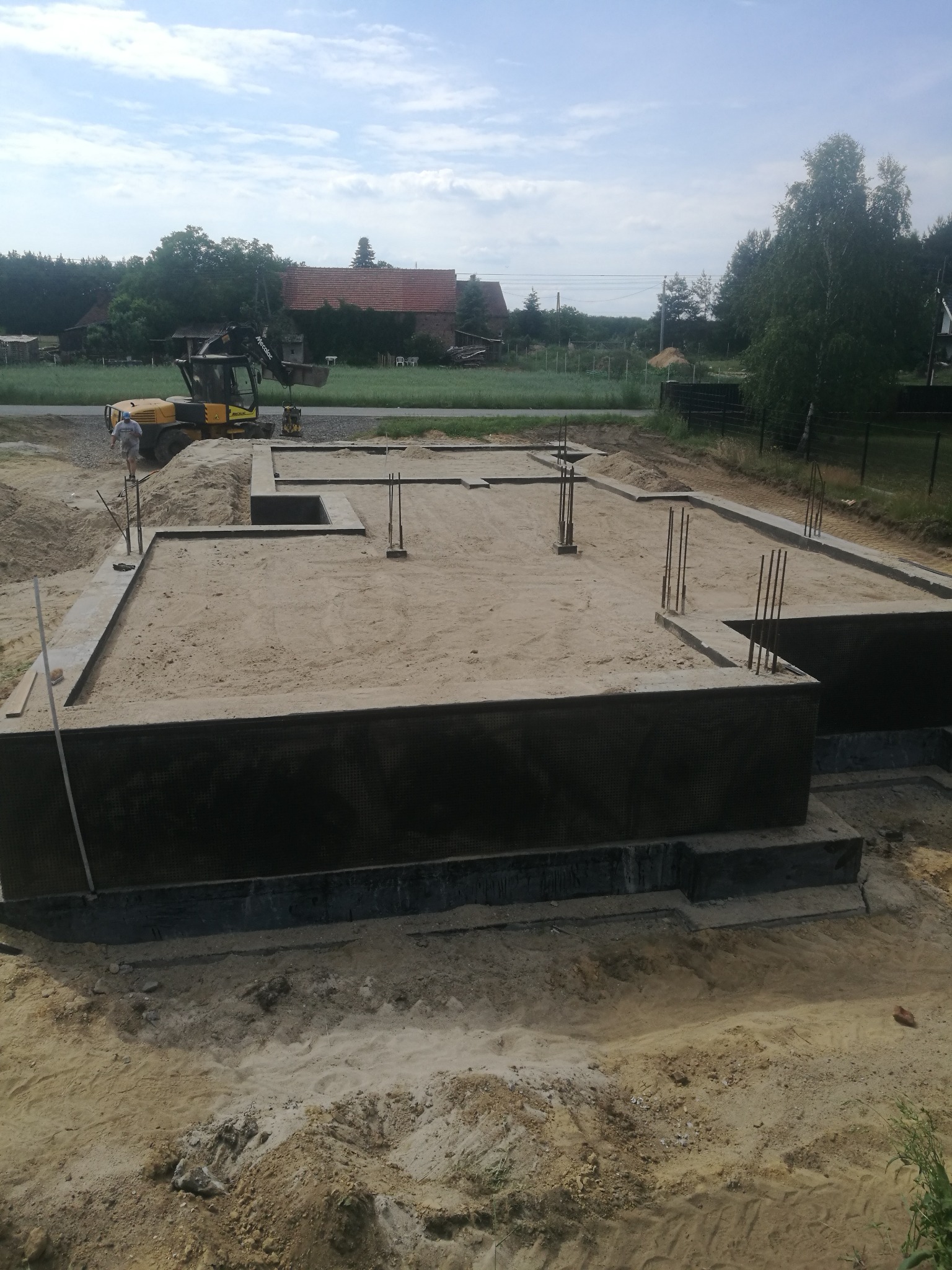 Zalane fundamenty domu z wystającymi prętami zbrojeniowymi, widoczny operator koparki i maszyna w tle, teren budowy w słoneczny dzień.
