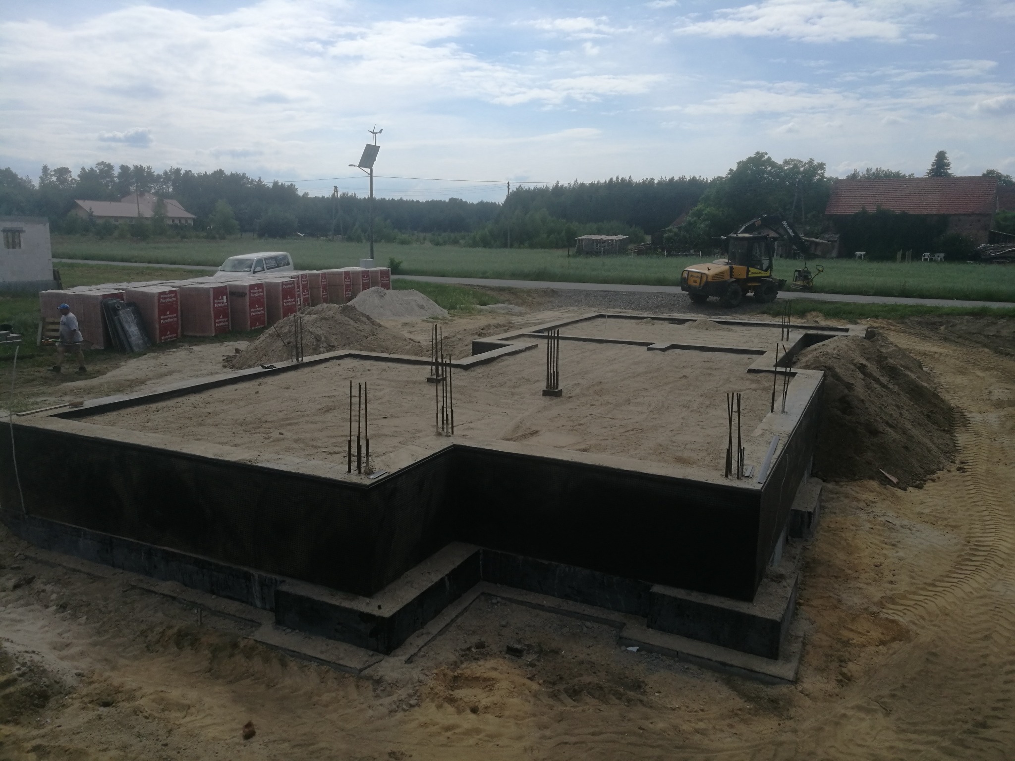 Wylany fundament domu z wystającymi prętami zbrojeniowymi, zabezpieczony czarną folią izolacyjną, z hałdą piasku i paletami cegieł Porotherm w tle, na działce budowlanej z widoczną koparką...