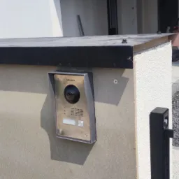 Zewnętrzny panel wideodomofonu Hikvision zamontowany na szarej, teksturowanej ścianie budynku z elementami drewnianymi i czarnymi detalami.