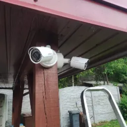 Dwie białe kamery monitoringu Dahua zamontowane pod zadaszeniem z brązowego drewna, widoczna drabina i śmietnik w tle.