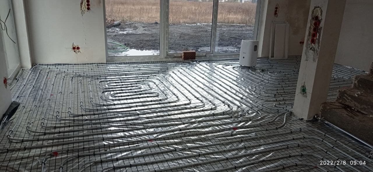 Szare rury ogrzewania podłogowego ułożone na metalowej siatce w surowym pomieszczeniu z widokiem na zewnątrz przez duże okno; widoczne niedokończone ściany i słupy.