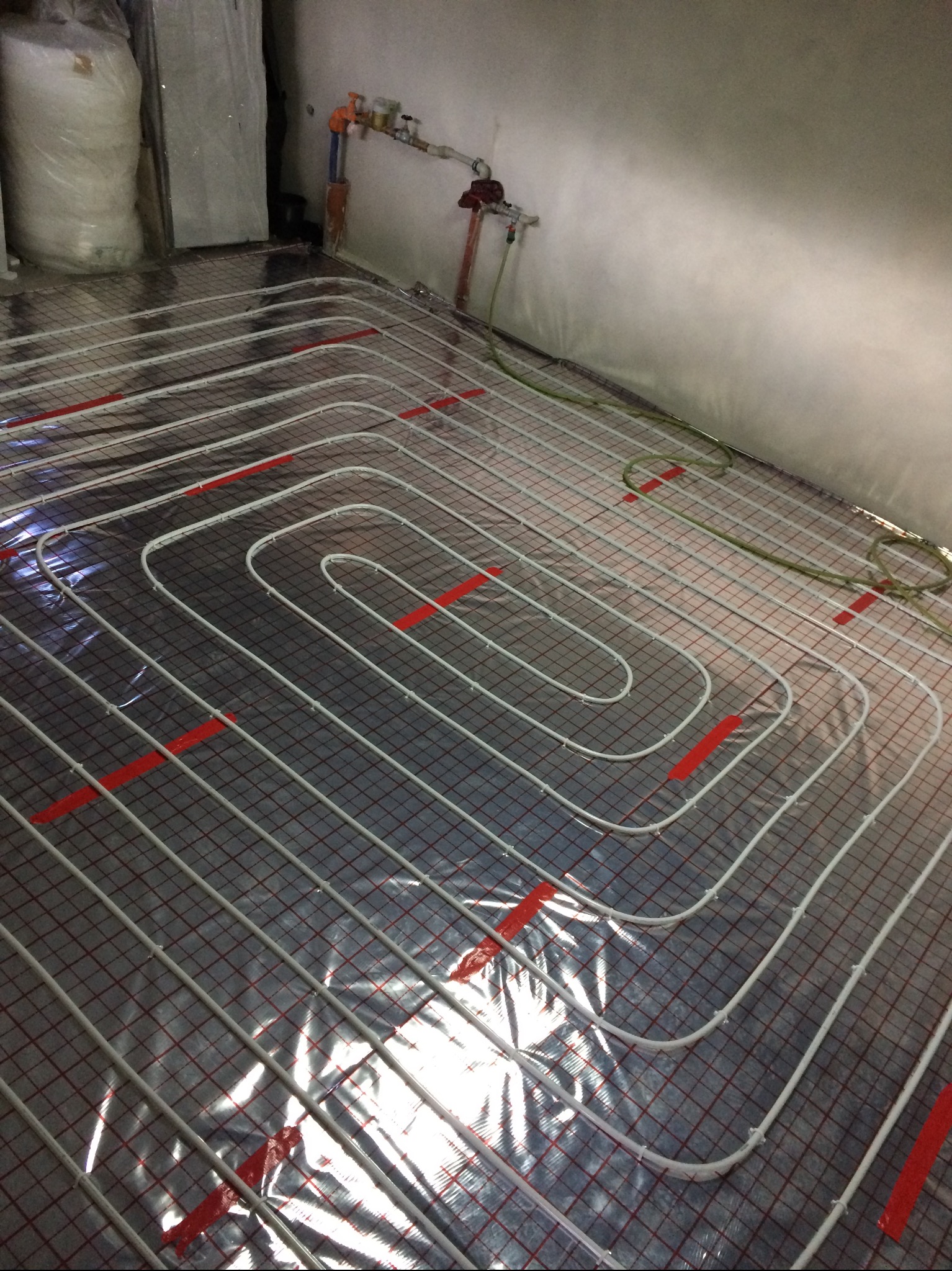 Białe rurki ogrzewania podłogowego ułożone na siatce z folii aluminiowej, przytwierdzone czerwoną taśmą, widoczne elementy instalacji na ścianie.