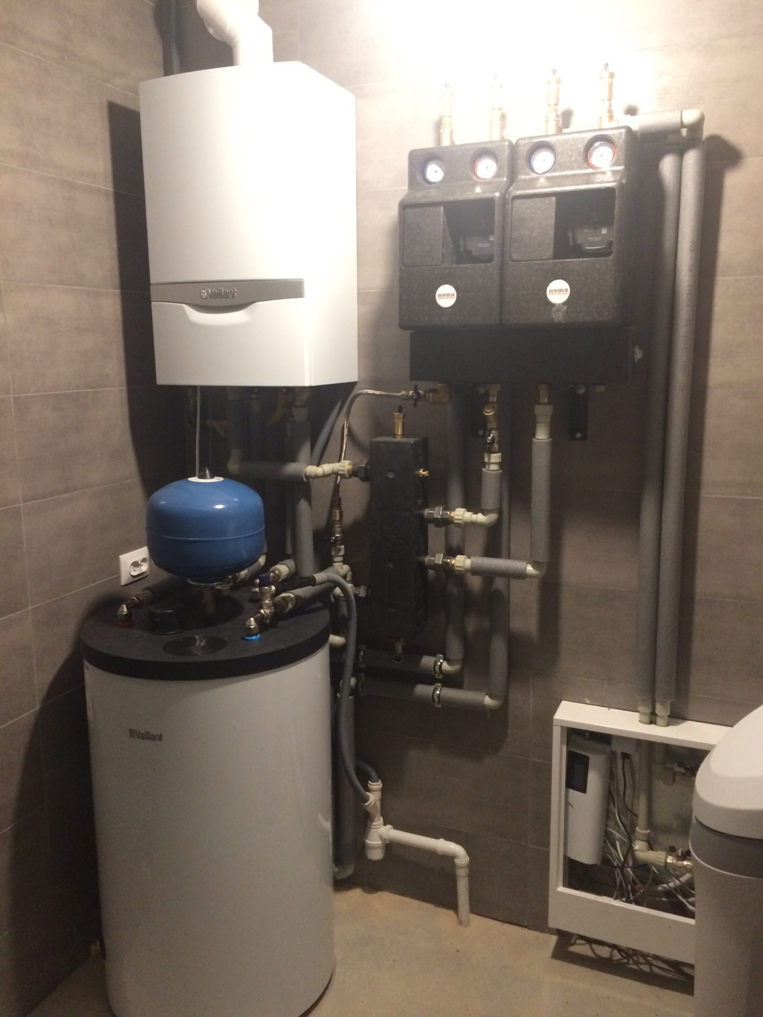 Zestaw urządzeń grzewczych marki Vaillant z niebieskim zbiornikiem wyrównawczym, czarnymi modułami rozdzielaczy z manometrami i szarymi rurami w kotłowni wykończonej płytkami w odcieniach szarości.