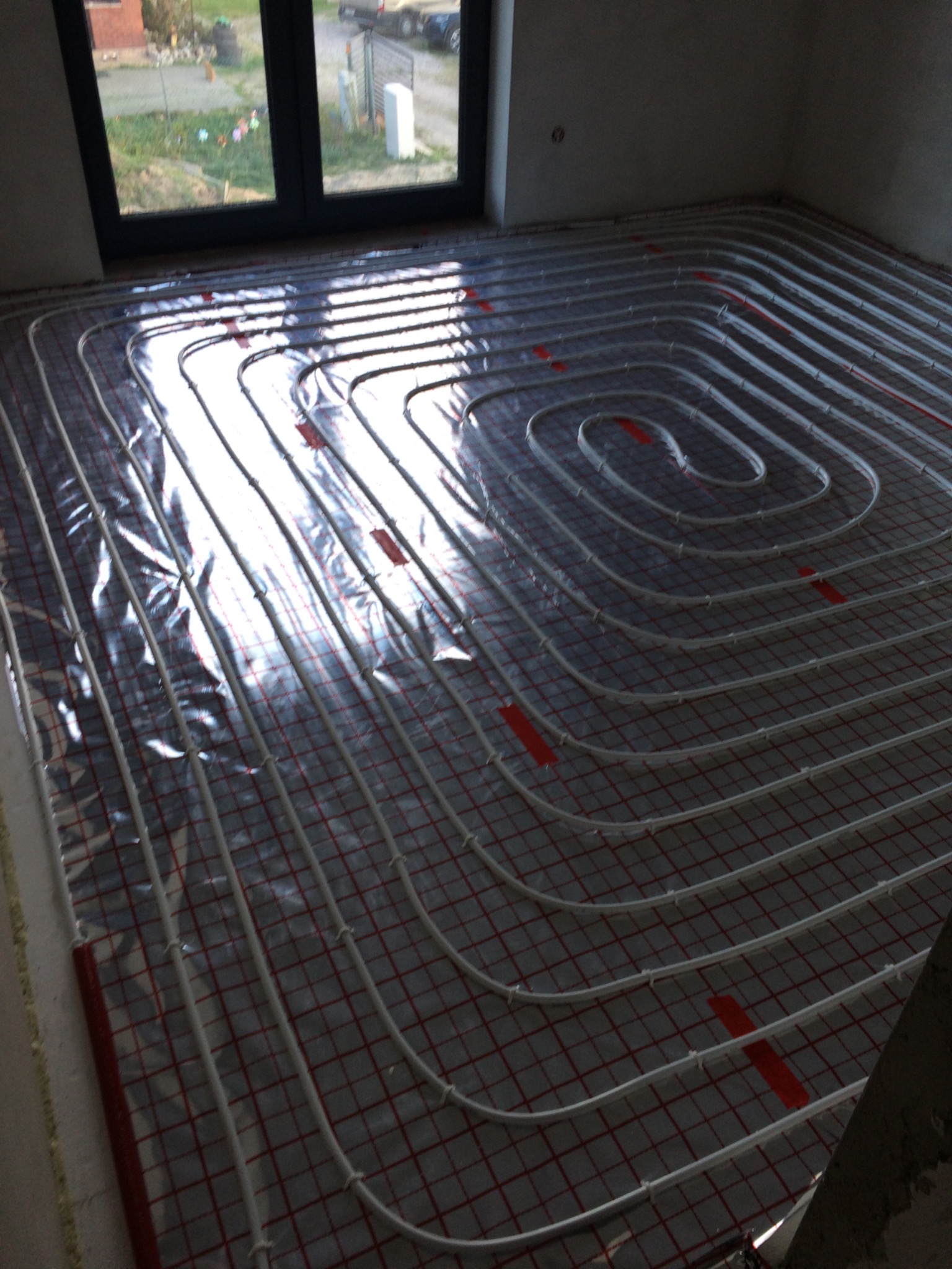 Rozłożona instalacja ogrzewania podłogowego w spiralnym układzie, rurki mocowane do siatki, widoczne okno w tle, surowy stan wykończenia.