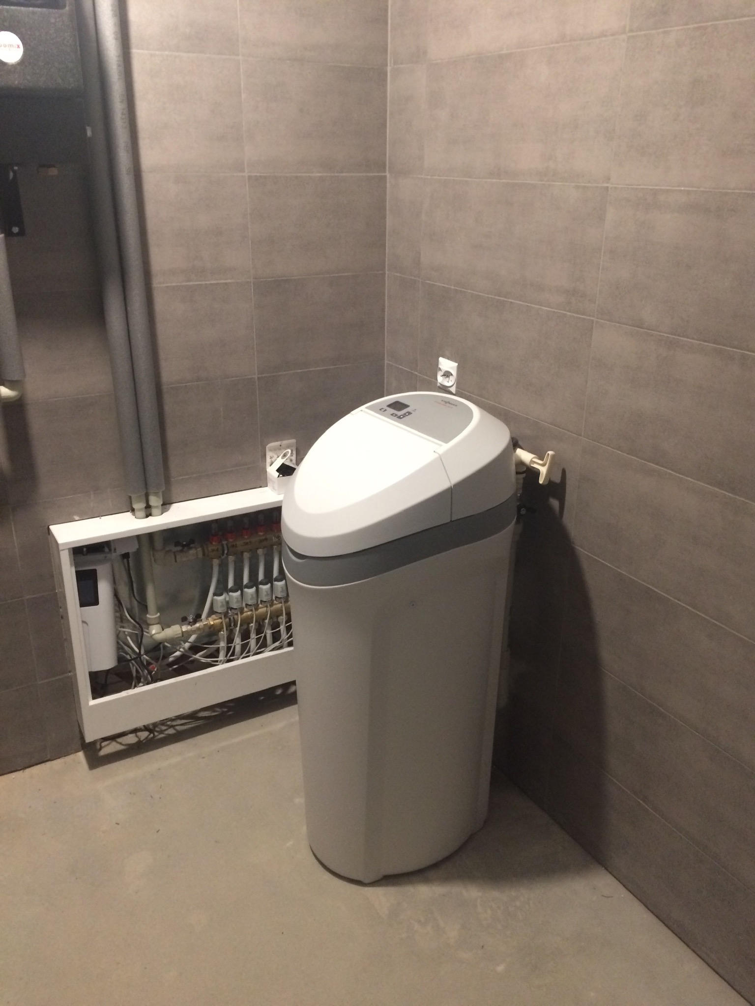 Szary zmiękczacz wody ustawiony obok białej szafki z instalacją grzewczą na tle szarych płytek ściennych, widoczna instalacja rurowa z zaworami.