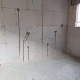 Wnętrze w stanie surowym z wyżłobieniami w ścianach pod instalację elektryczną; widoczne pionowe bruzdy i okrągłe otwory w białych bloczkach ścian.