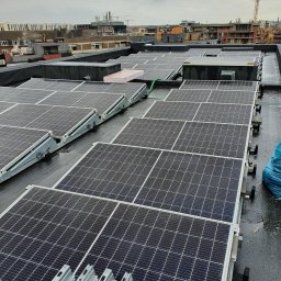 Firma Usługowa Work Service - Rząd paneli słonecznych na płaskim dachu budynku miejskiego w pochmurny dzień, widoczne elementy montażowe i drabina w rogu.