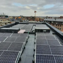 Widok z góry na panele słoneczne zamontowane na płaskim dachu budynku miejskiego w pochmurny dzień, widoczna instalacja elektryczna i elementy mocujące.