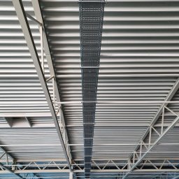 Firma Usługowa Work Service - Metalowa korytka kablowa podwieszona pod sufitem hali przemysłowej, widoczne elementy konstrukcji stalowej dachu i blaszane poszycie sufitu.