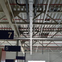 Firma Usługowa Work Service - Metalowa konstrukcja sufitowa z instalacjami elektrycznymi, rurami i kanałami wentylacyjnymi w hali magazynowej, widok z dołu.