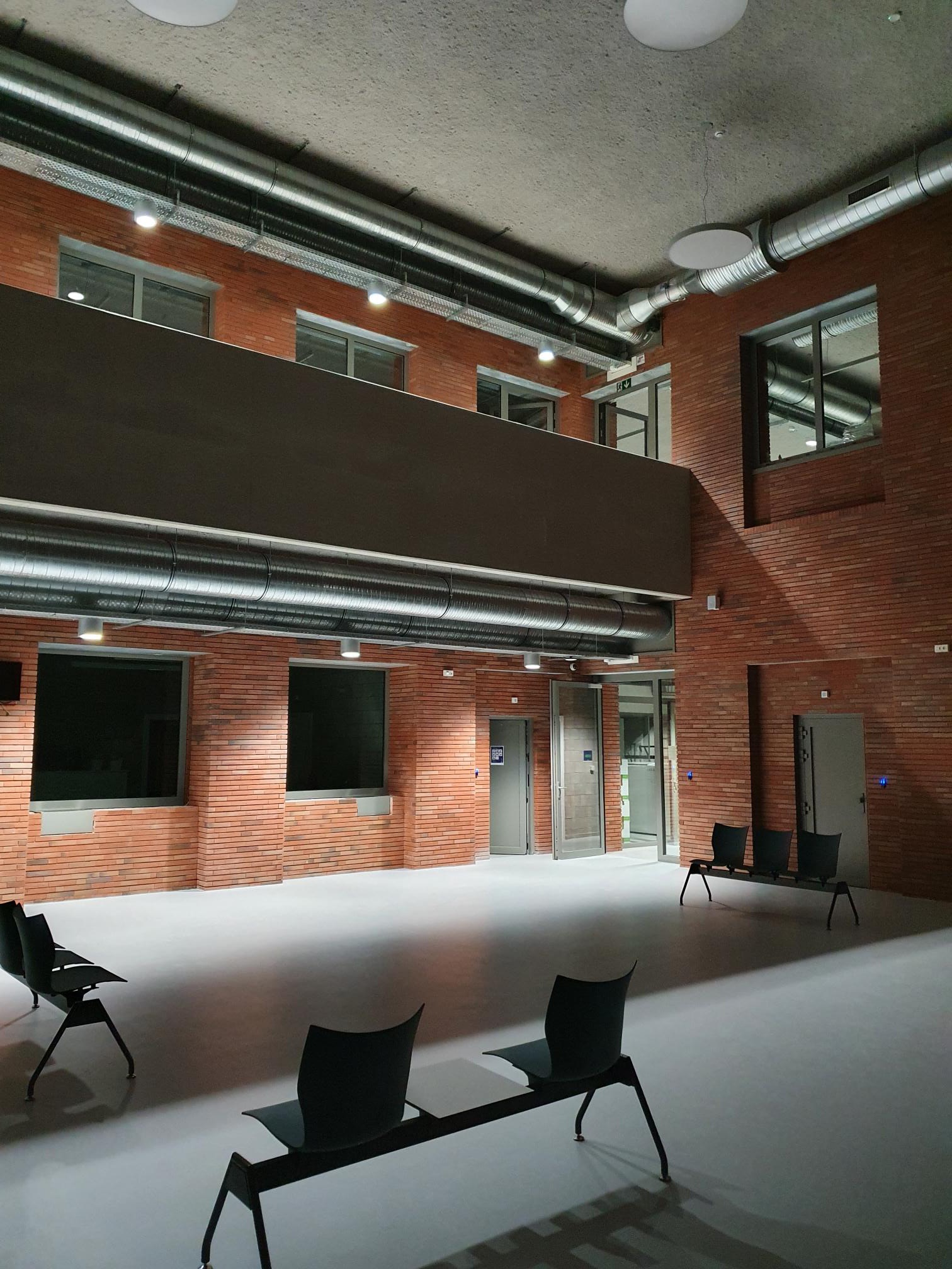 Wnętrze budynku z widocznymi instalacjami wentylacyjnymi pod sufitem, oświetleniem punktowym i ścianami z cegły, z czarnymi ławkami dla oczekujących.