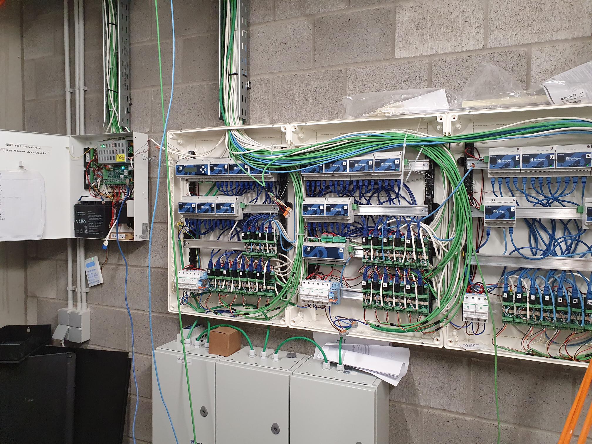 Rozdzielnica alarmowa z licznymi modułami Syntegro, okablowaniem w kolorach zielonym i niebieskim, zamontowana na szarej ścianie z betonowych bloczków, widoczne także inne elementy instalacji...