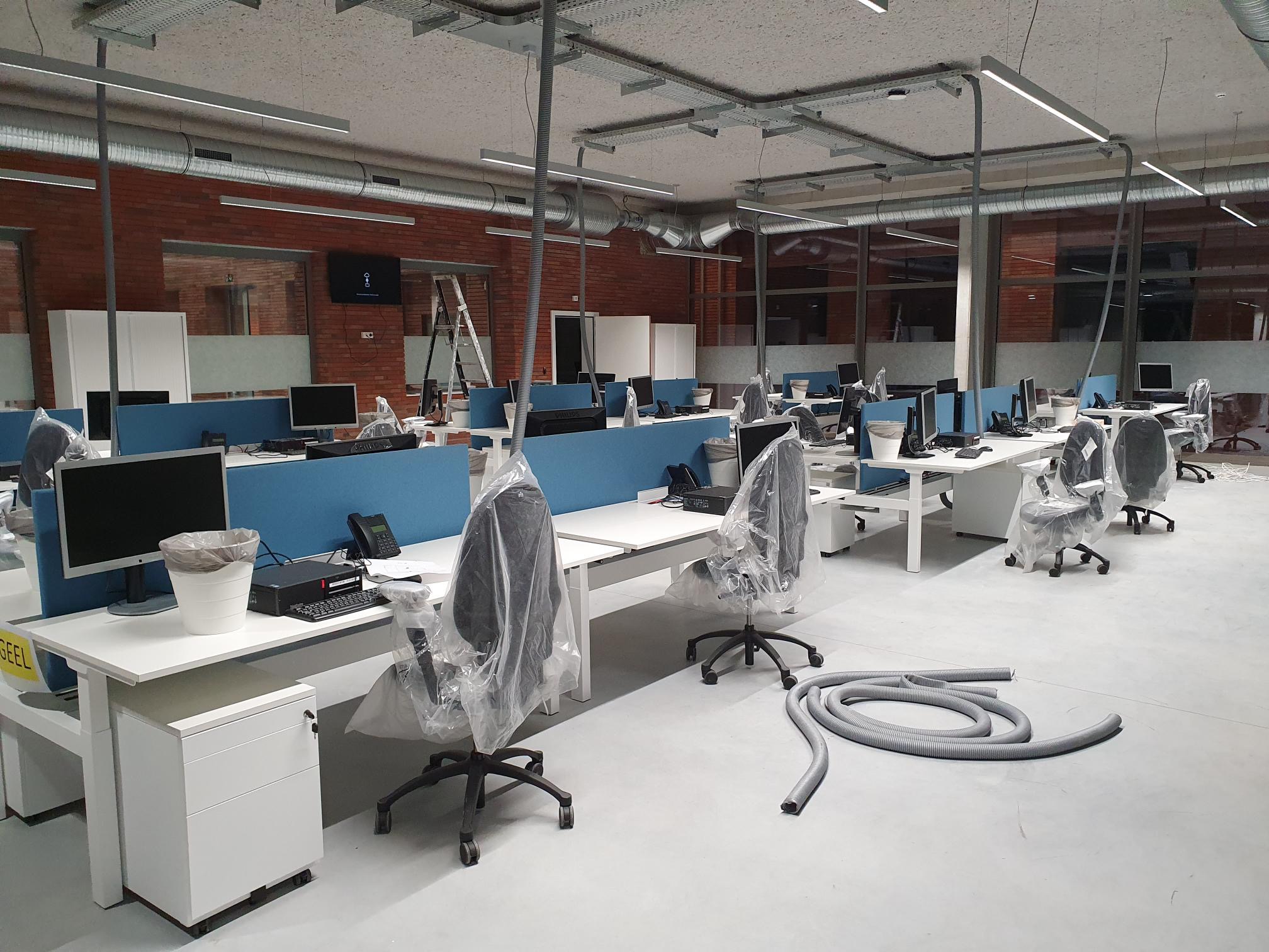 Nowe biuro w trakcie przygotowań: białe biurka z niebieskimi przegrodami, krzesła zabezpieczone folią, widoczne instalacje pod sufitem i fragmenty przewodów wentylacyjnych na podłodze, ściana...