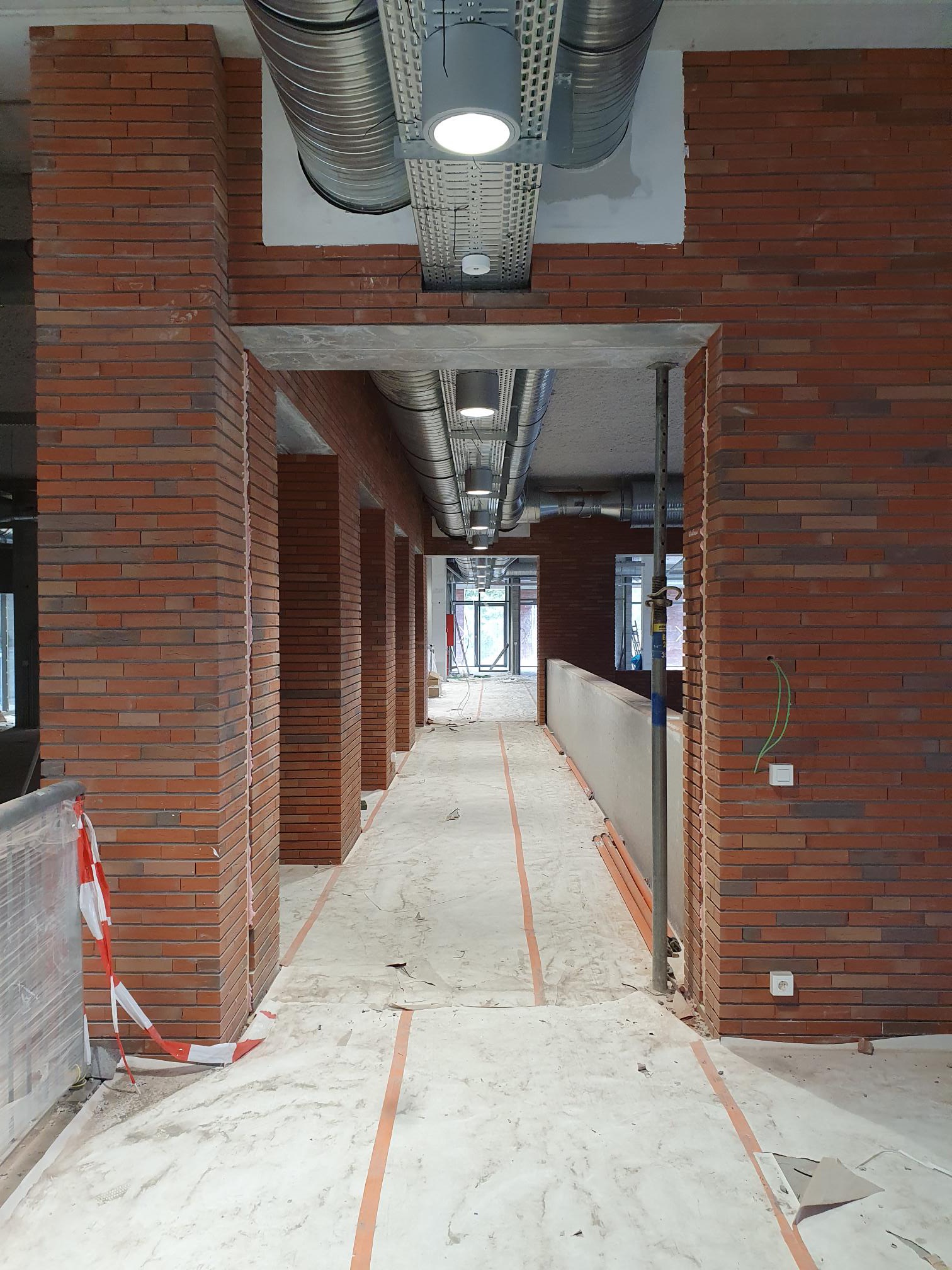 Wnętrze budynku w trakcie wykończenia z widocznymi instalacjami pod sufitem i fragmentami muru z cegły klinkierowej, zabezpieczone taśmą ostrzegawczą.