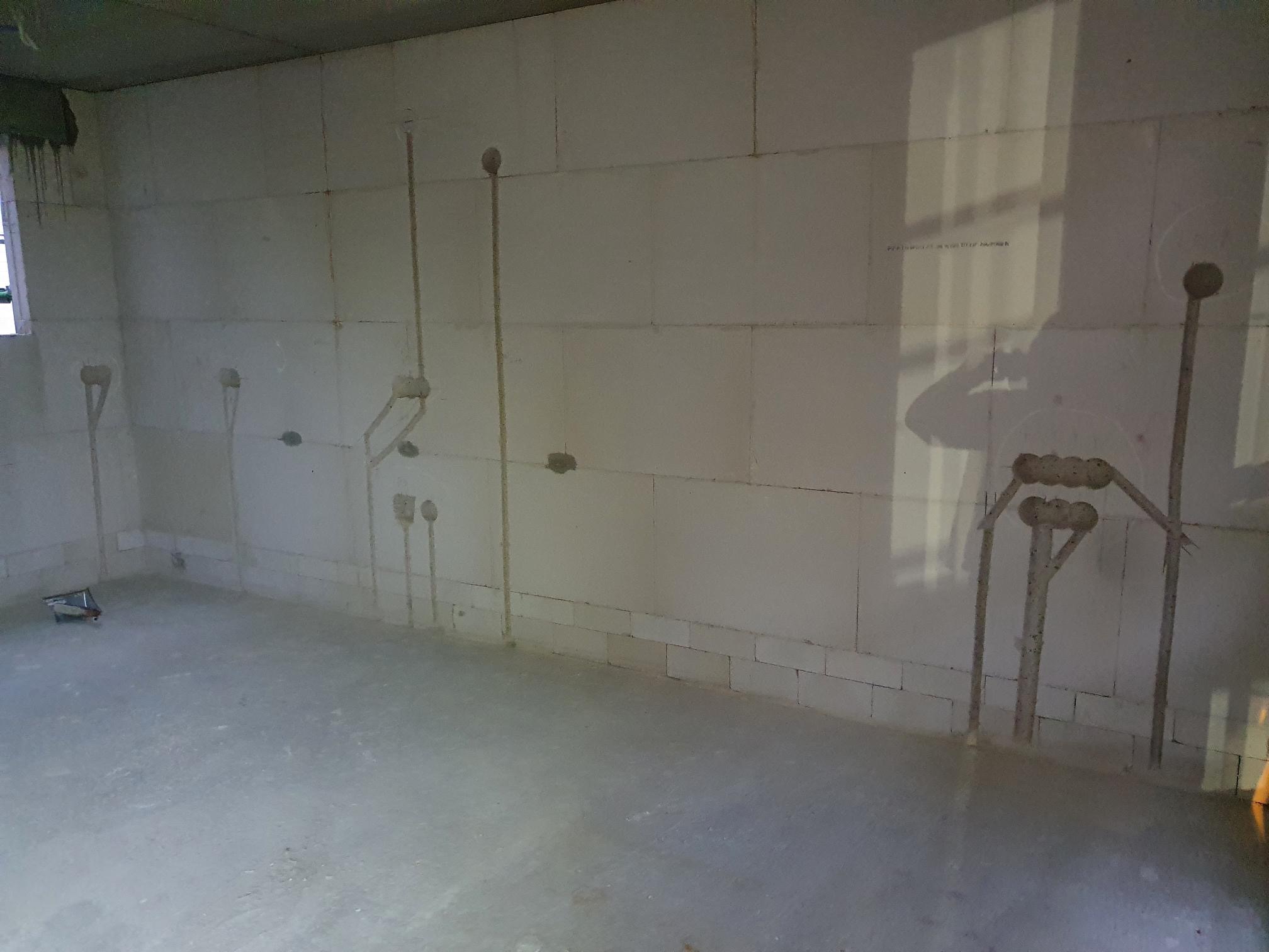 Surowy stan pomieszczenia z białymi ścianami z betonu komórkowego, widoczne pionowe rury instalacji elektrycznej wystające ze ścian, przygotowane pod gniazdka i oświetlenie.