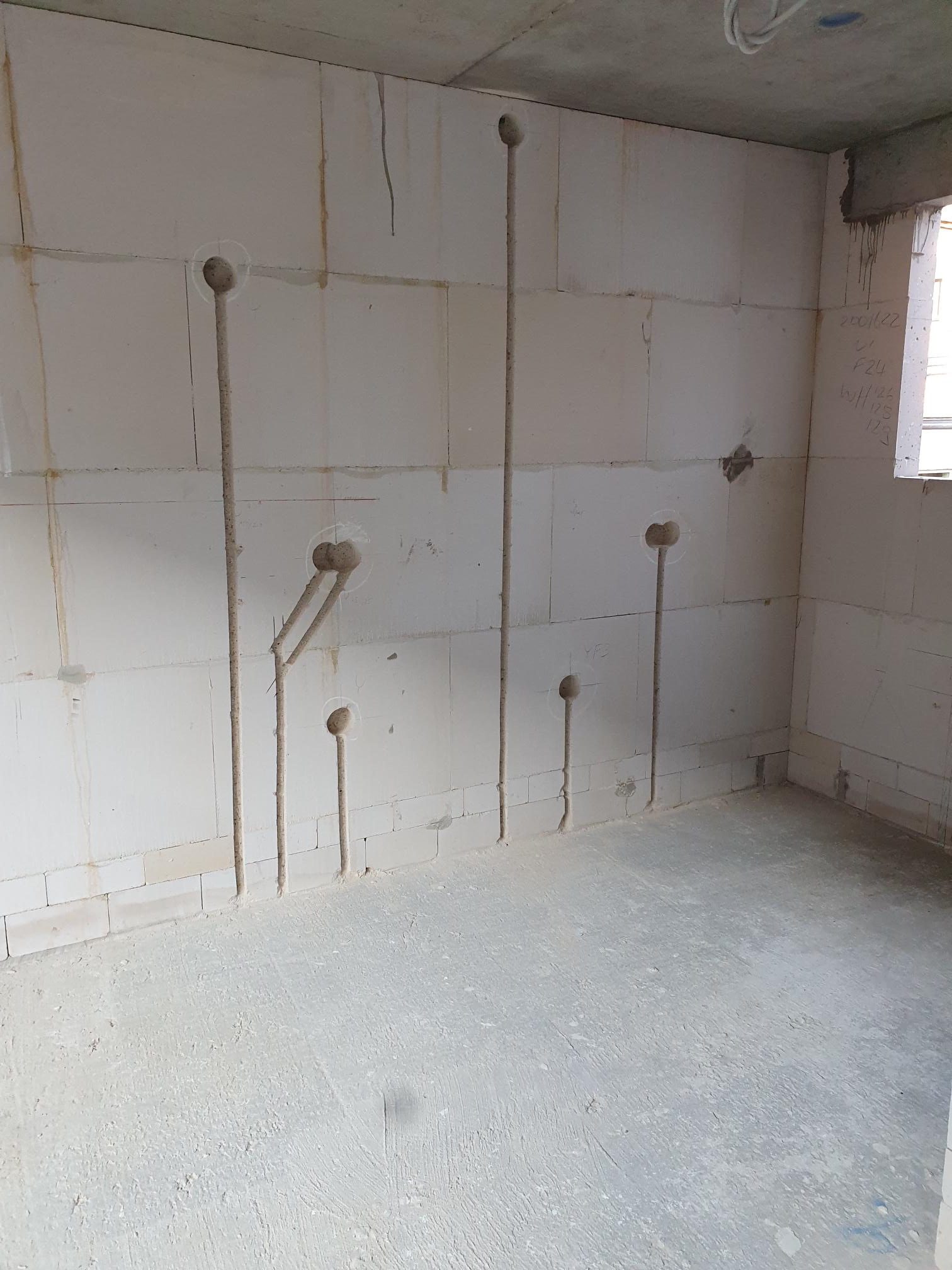 Wnętrze w stanie surowym z wyżłobieniami w ścianach pod instalację elektryczną; widoczne pionowe bruzdy i okrągłe otwory w białych bloczkach ścian.