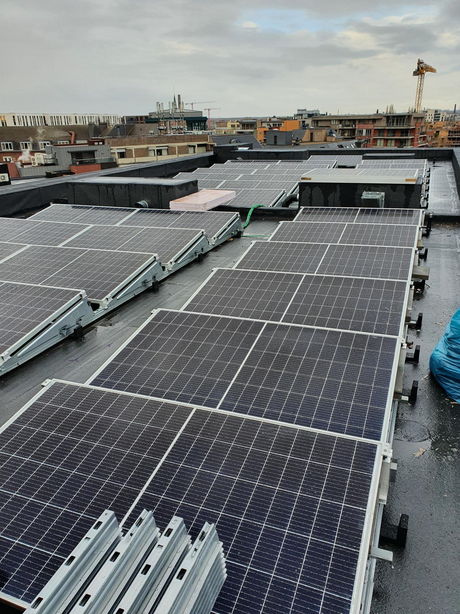 Rząd paneli słonecznych na płaskim dachu budynku miejskiego w pochmurny dzień, widoczne elementy montażowe i drabina w rogu.