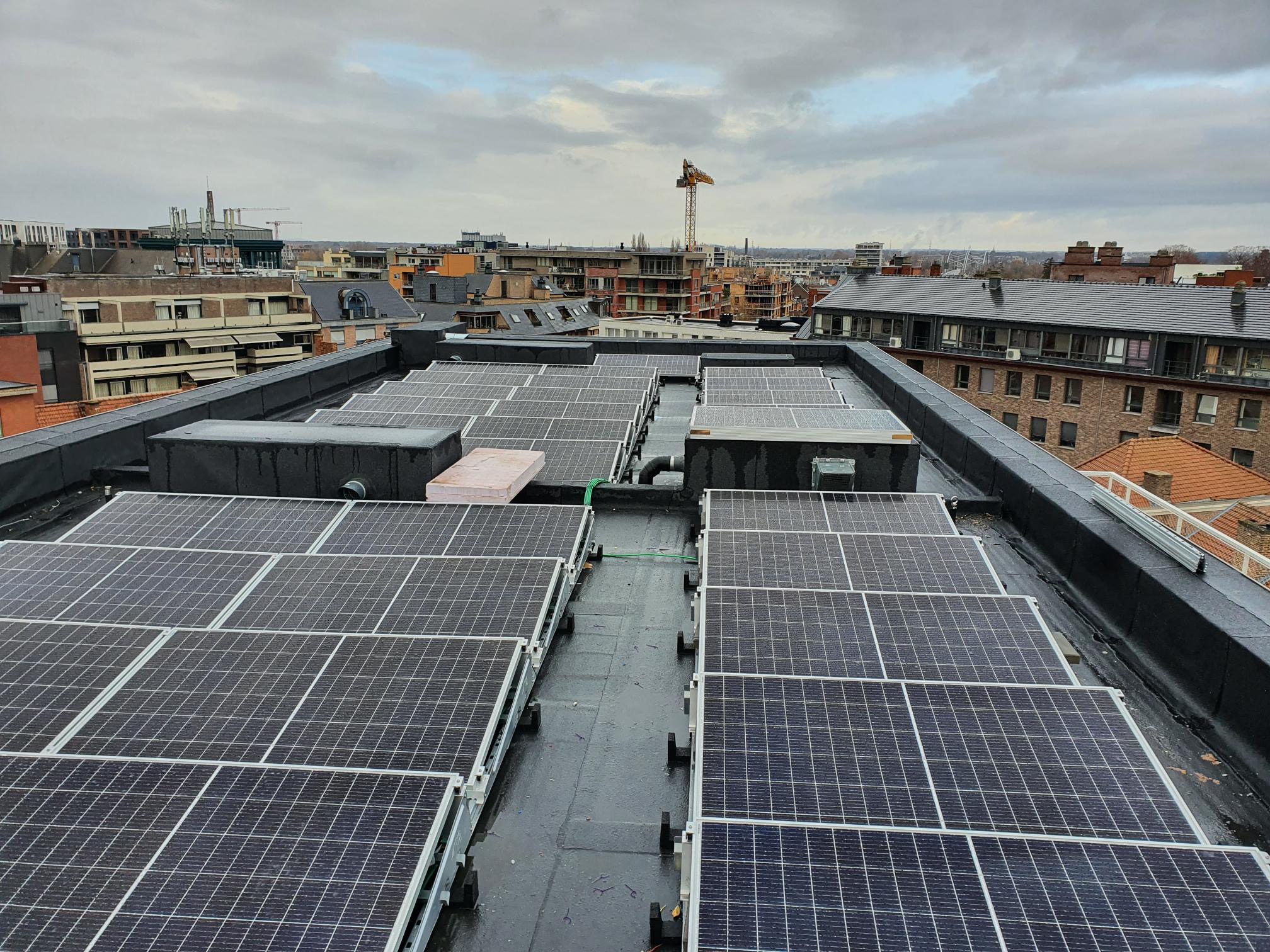 Widok z góry na panele słoneczne zamontowane na płaskim dachu budynku miejskiego w pochmurny dzień, widoczna instalacja elektryczna i elementy mocujące.