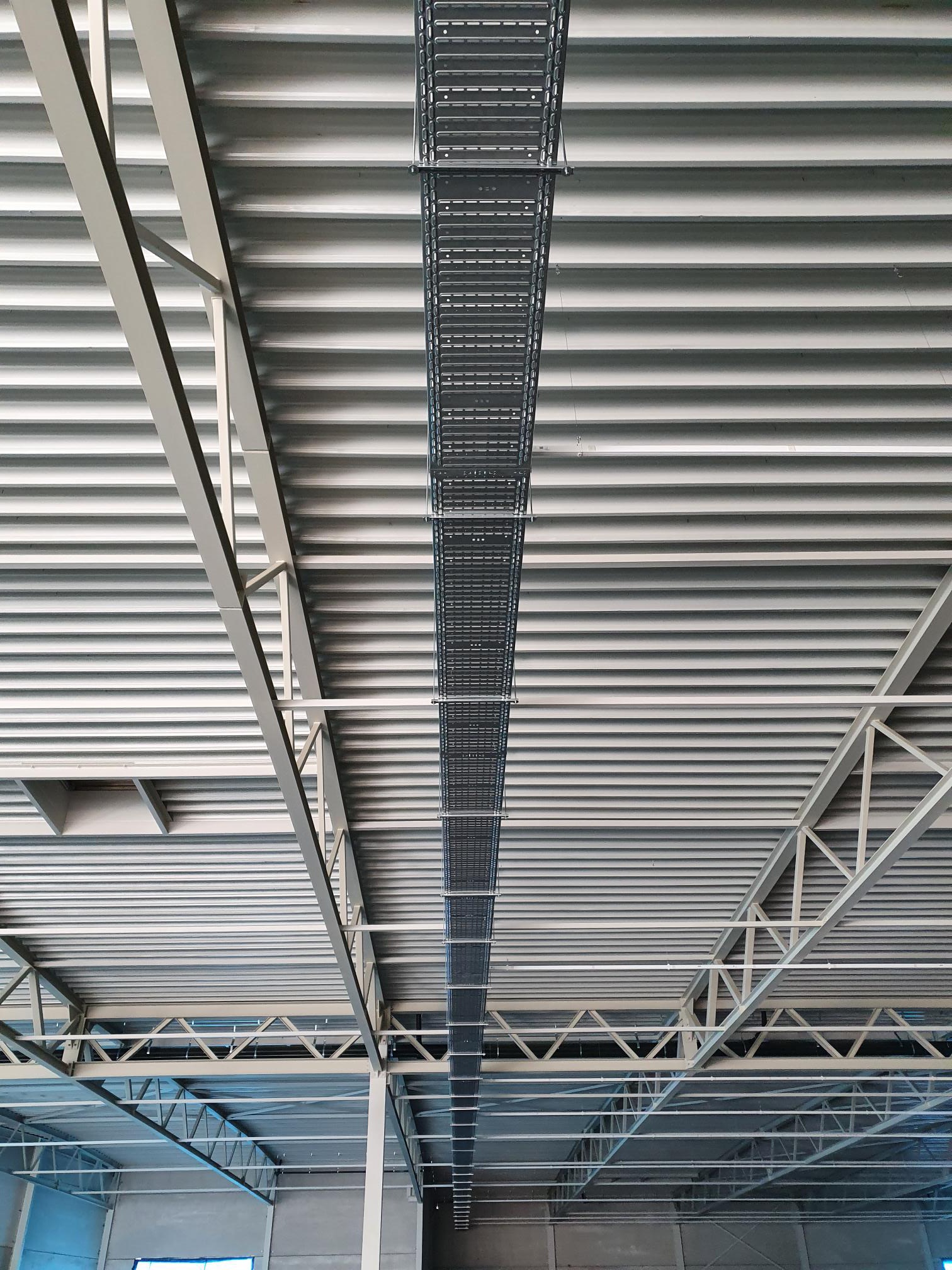 Metalowa korytka kablowa podwieszona pod sufitem hali przemysłowej, widoczne elementy konstrukcji stalowej dachu i blaszane poszycie sufitu.
