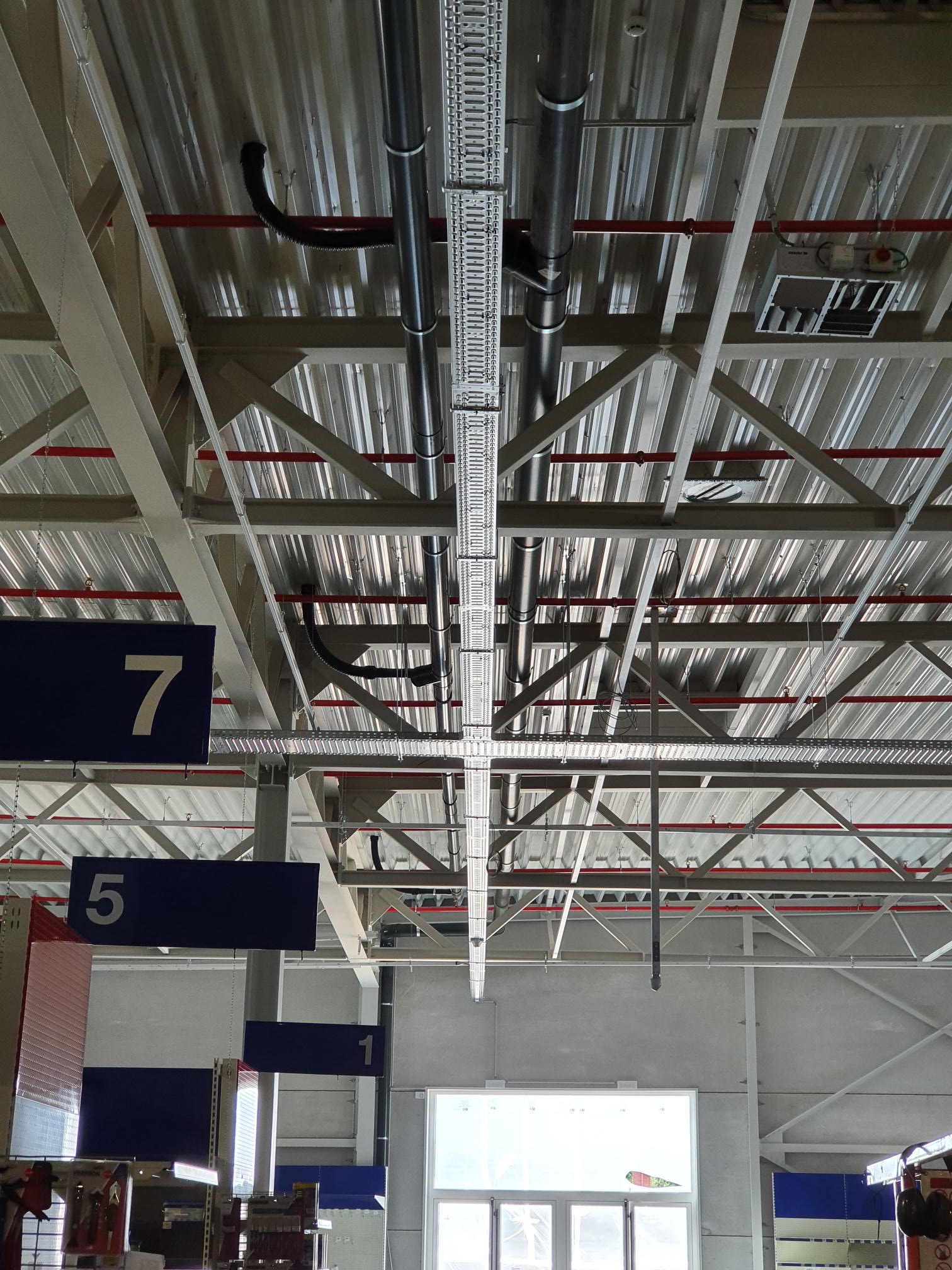 Metalowa konstrukcja sufitowa z instalacjami elektrycznymi, rurami i kanałami wentylacyjnymi w hali magazynowej, widok z dołu.
