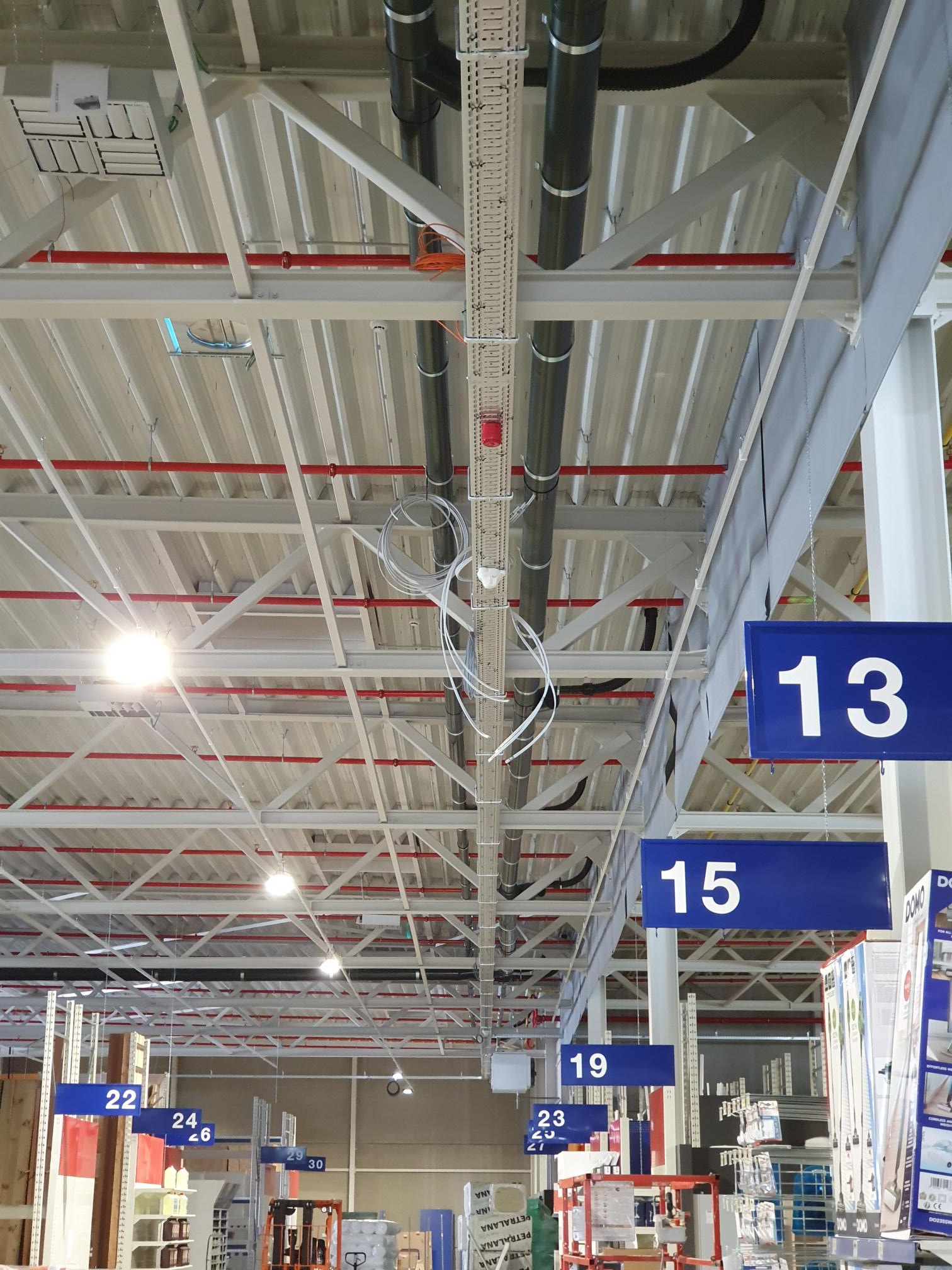 Sufit hali magazynowej z widocznymi elementami instalacji: rury, korytka kablowe z przewodami, system przeciwpożarowy z czerwoną głowicą tryskacza, oraz oznaczenia numerów alejek w postaci...