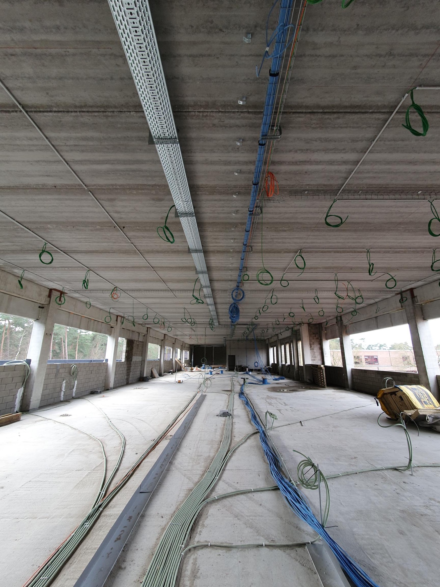 Wnętrze hali w trakcie instalacji elektrycznej: koryta kablowe pod sufitem, wiązki przewodów na podłodze, widoczne okna i surowa konstrukcja budynku.