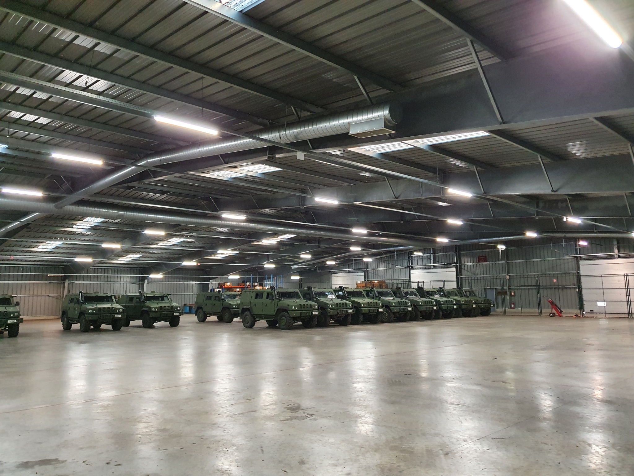 Rząd zaparkowanych, wojskowych pojazdów wewnątrz dużego, oświetlonego hali magazynowej z metalową konstrukcją dachu.