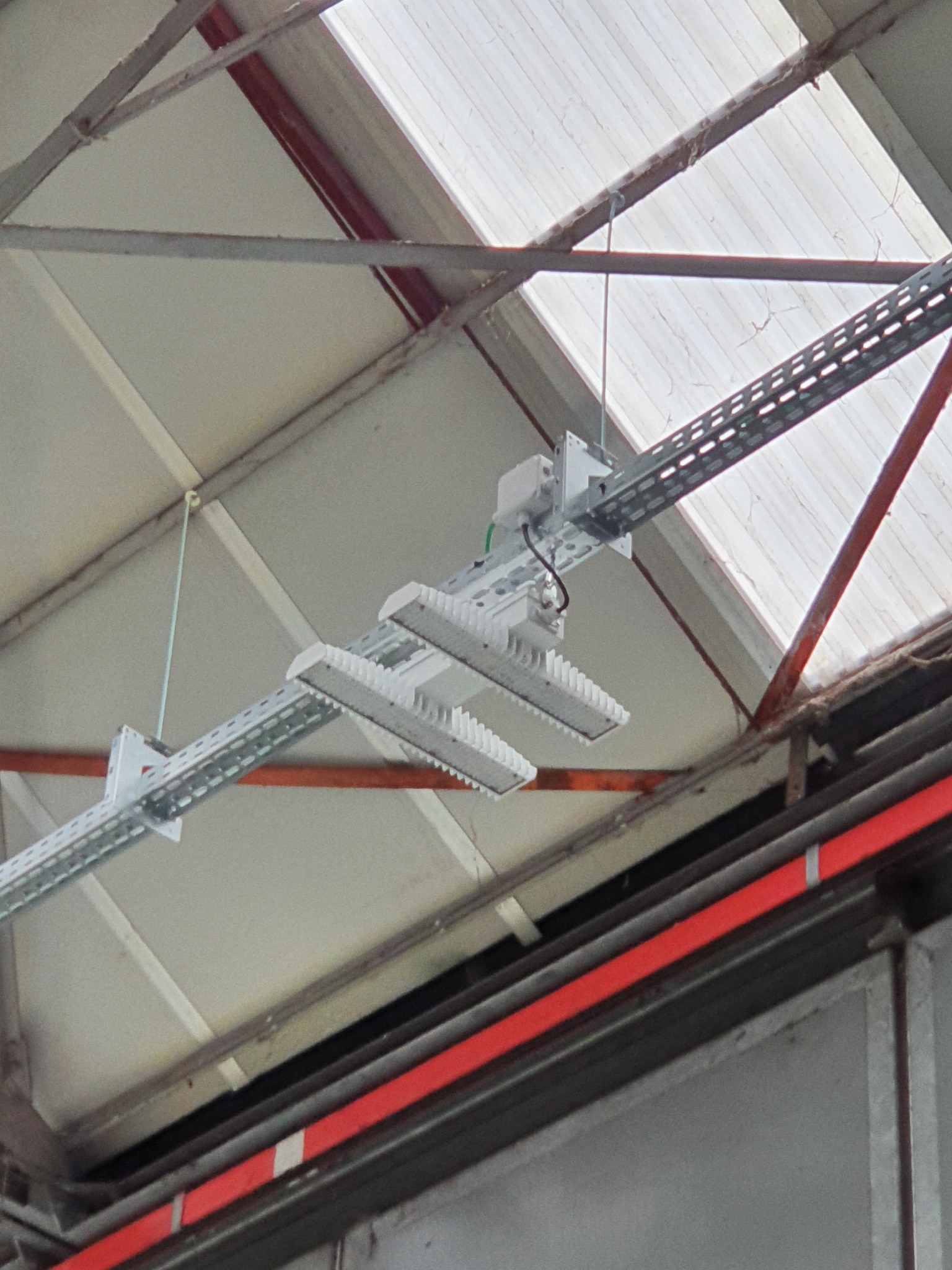 Zamontowane, podwójne lampy LED w metalowych korytkach kablowych, zawieszone pod sufitem hali z widoczną konstrukcją stalową i świetlikiem.