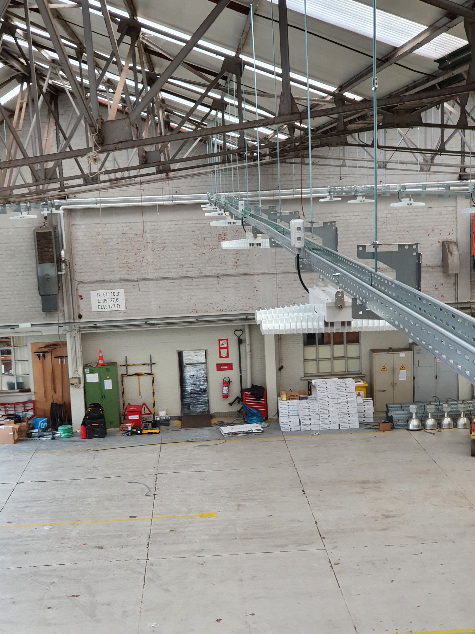 Wnętrze hali przemysłowej z zamontowanymi szynami oświetleniowymi, widoczne lampy, elementy konstrukcyjne dachu, sprzęt i narzędzia na podłodze.