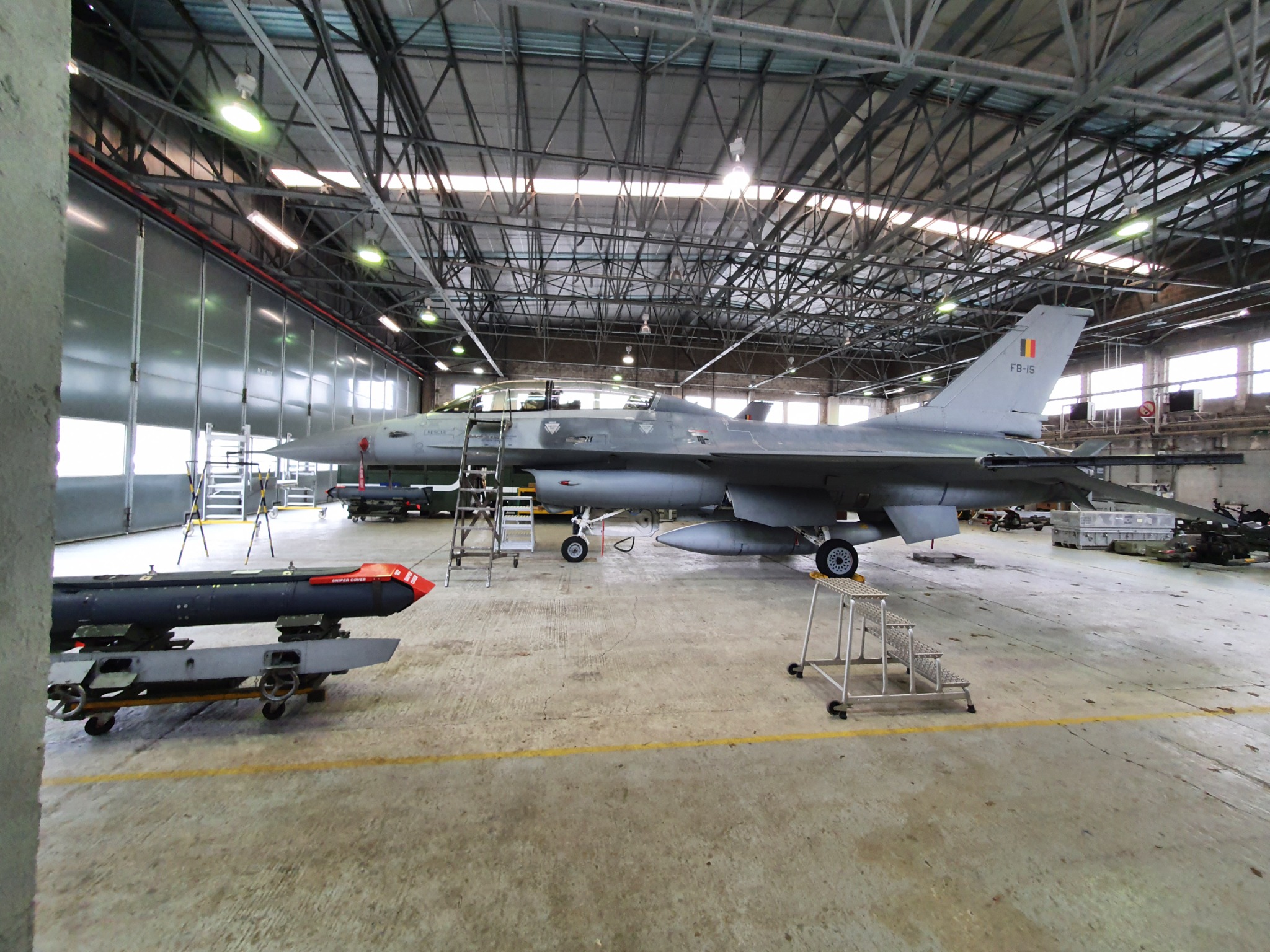 Myśliwiec F-16 z flagą Belgii na stateczniku pionowym, zaparkowany w hangarze z widocznymi rampami i amunicją na wózkach.