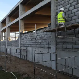 Pracownik w kasku i kamizelce na rusztowaniu, muruje ścianę z szarego betonu komórkowego na konstrukcji żelbetowej z drewnianym stropem.