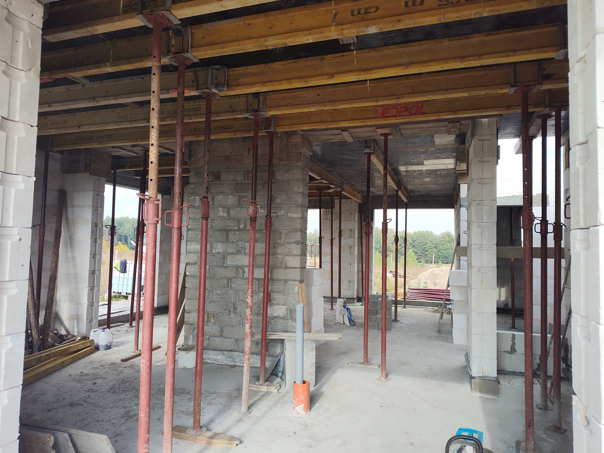 Wnętrze surowego domu w trakcie budowy, widoczne podpory stropowe, ściany wymurowane z białych bloczków, szalunki stropowe z belek drewnianych, widok na zewnątrz przez otwory okienne.