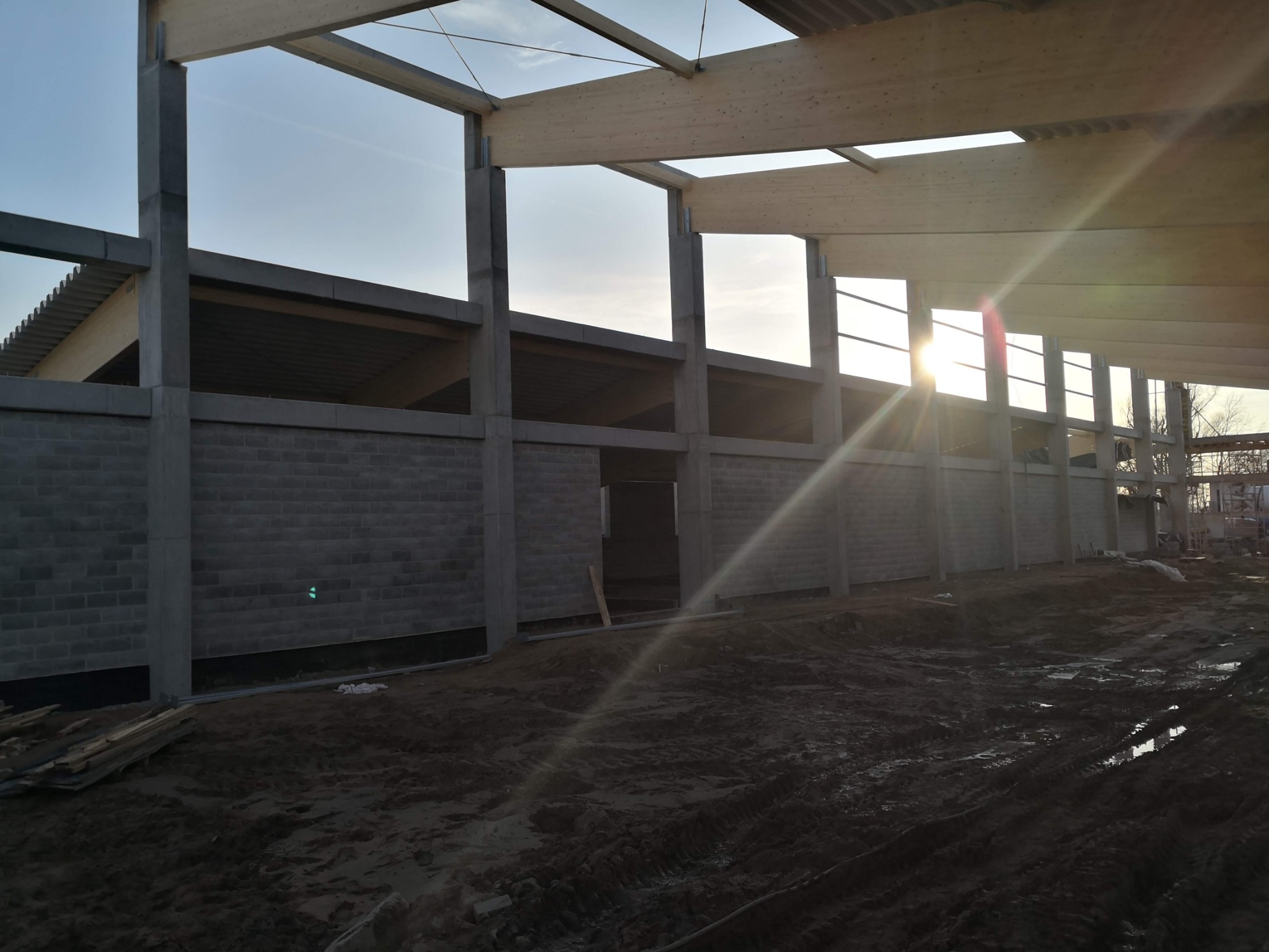 Surowy stan budowy: betonowe ściany z pustaków i drewniana konstrukcja dachu w świetle zachodzącego słońca, ziemiste podłoże.