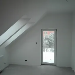 Poddasze z oknem balkonowym, widok na zaśnieżony ogród, białe ściany i grzejnik, surowy beton na podłodze, widoczne elementy konstrukcyjne dachu.