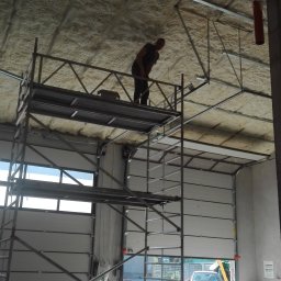 Sebastian Góralczyk - Pracownik na rusztowaniu aplikuje pianę izolacyjną na suficie wewnątrz budynku z częściowo otwartą bramą garażową.