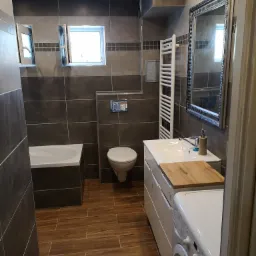 Nowoczesna łazienka z brązowymi płytkami na ścianach i podłodze, białą armaturą, drewnianym blatem przy umywalce i pralką widoczną w kadrze.