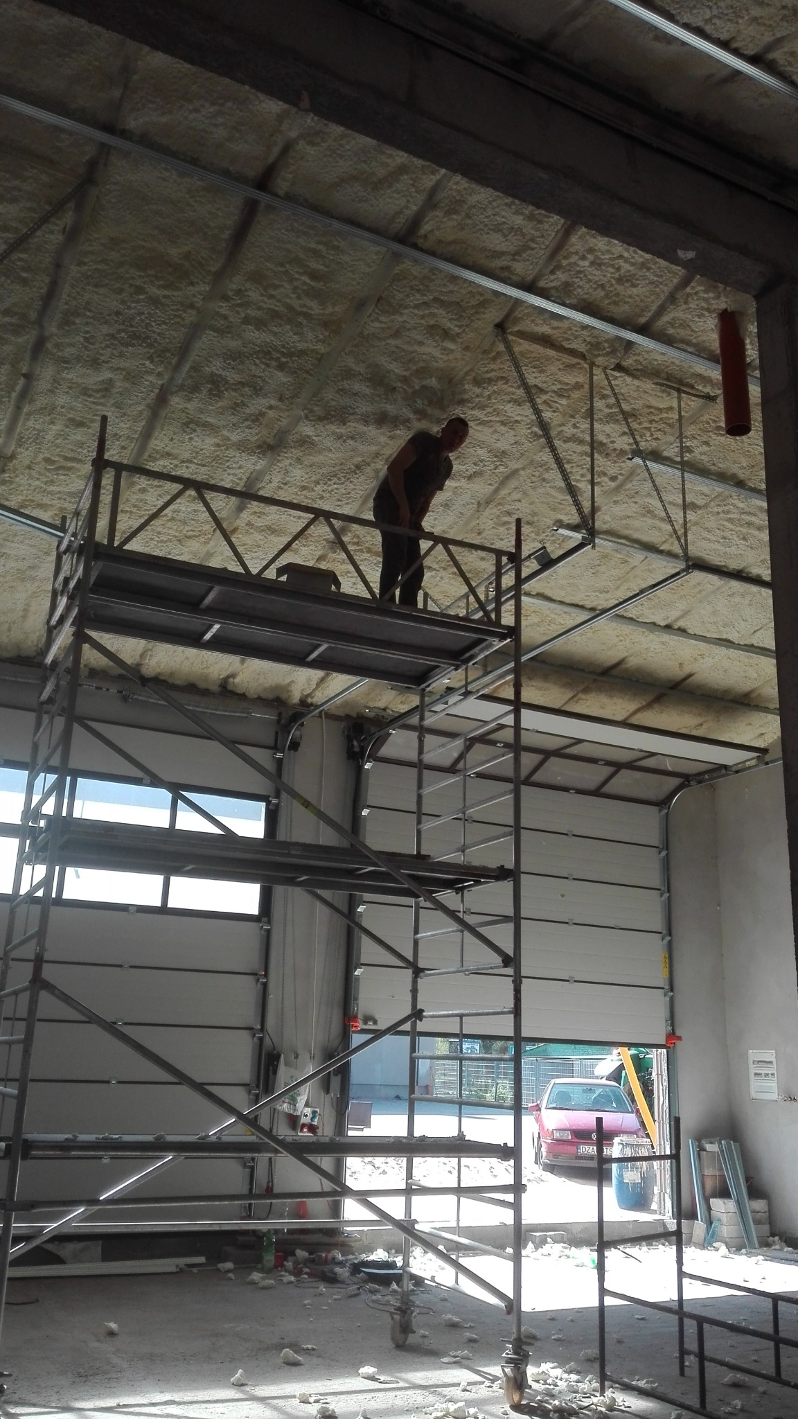 Pracownik na rusztowaniu aplikuje pianę izolacyjną na suficie wewnątrz budynku z częściowo otwartą bramą garażową.