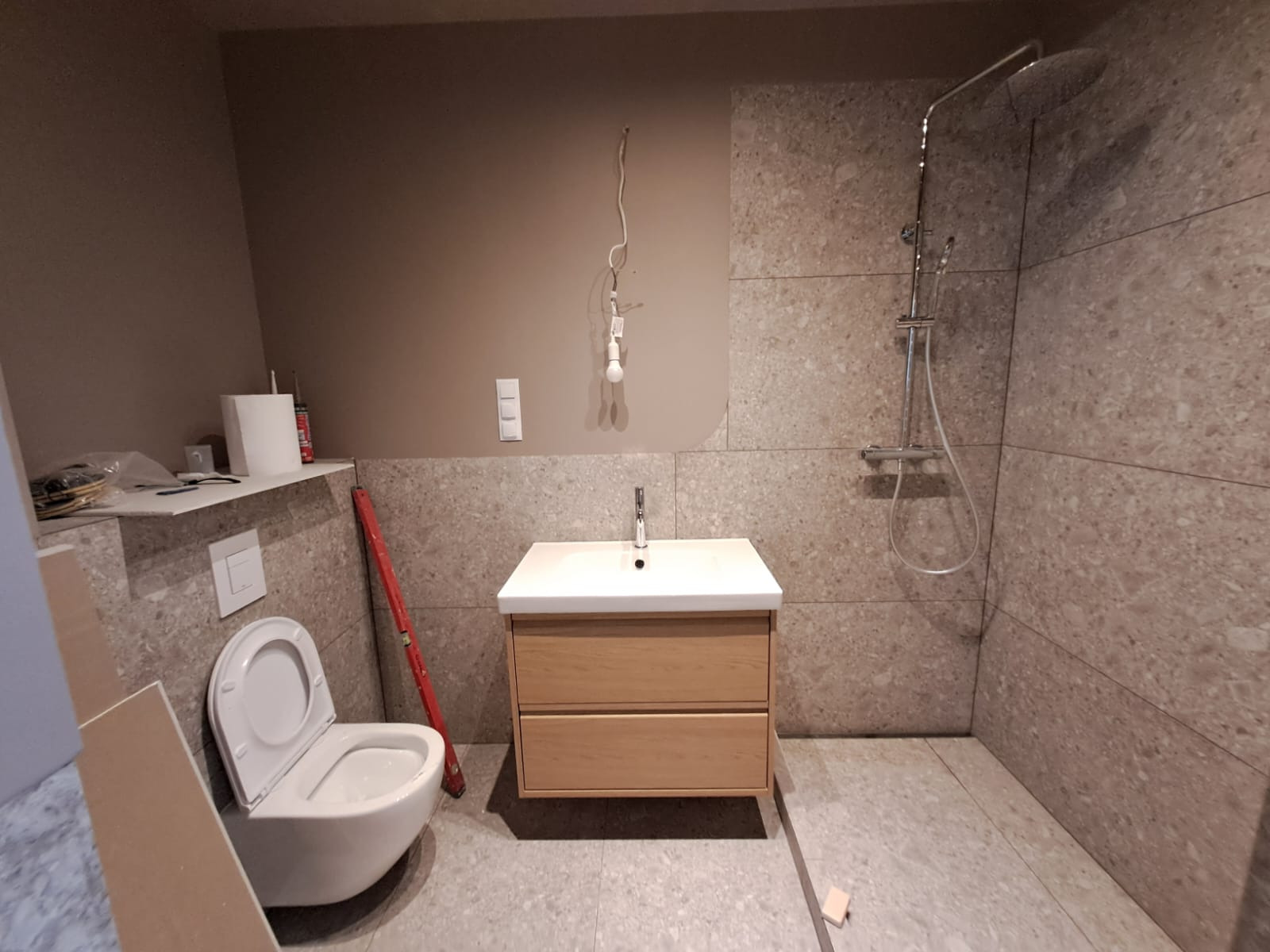 Nowoczesna łazienka w trakcie remontu: biała umywalka z drewnianą szafką, otwarta toaleta, szara mozaika na ścianach i podłodze, widoczne narzędzia i niedokończone oświetlenie.