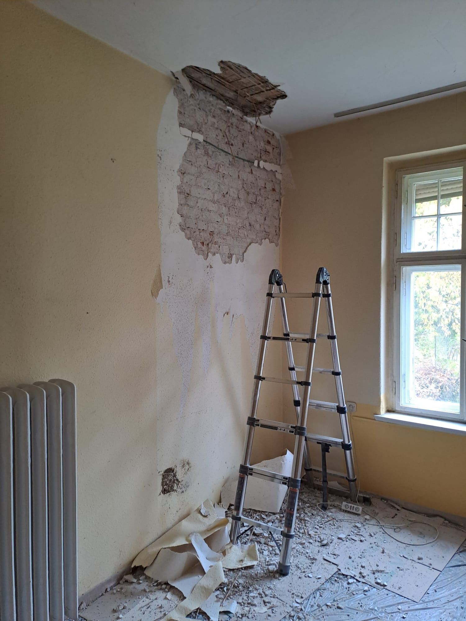 Pokój w trakcie remontu: częściowo zerwana tapeta, odsłonięta cegła na ścianie i dziura w suficie. Aluminiowa drabina stoi na podłodze pokrytej resztkami materiałów.