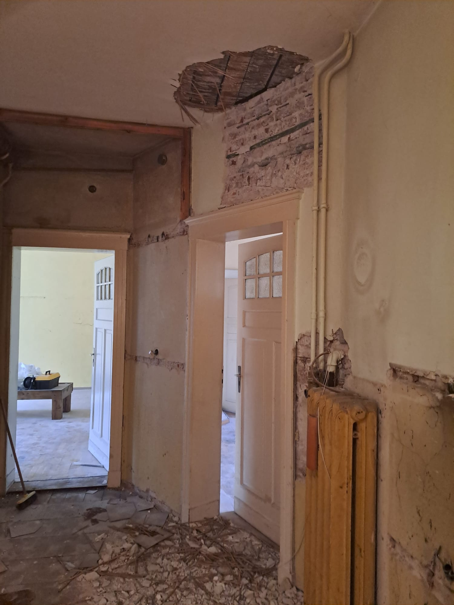 Wnętrze mieszkania w trakcie remontu: odsłonięta cegła, dziura w suficie, stara instalacja grzewcza, widoczne ślady demontażu. Podłoga pokryta gruzem i resztkami materiałów.
