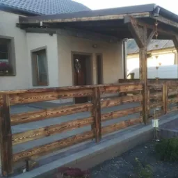 Zadaszony taras z drewnianą balustradą, widoczne detale konstrukcji dachu i impregnacja drewna, w tle budynek mieszkalny i samochód dostawczy.