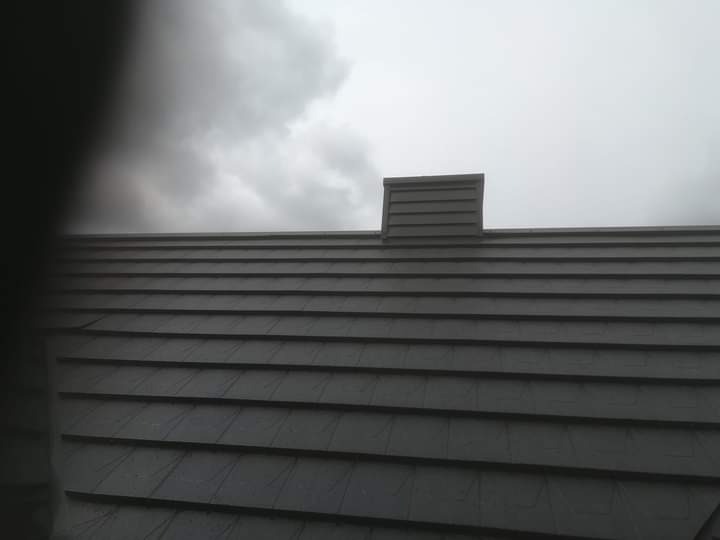 Szary dach pokryty dachówką bitumiczną z kominkiem wentylacyjnym na tle pochmurnego nieba.