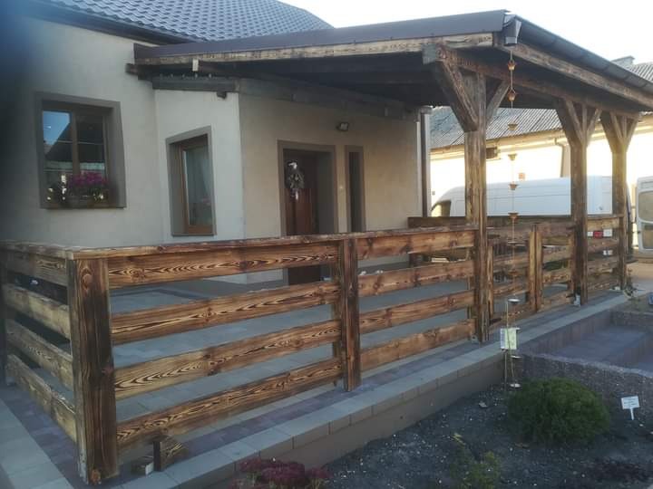 Zadaszony taras z drewnianą balustradą, widoczne detale konstrukcji dachu i impregnacja drewna, w tle budynek mieszkalny i samochód dostawczy.