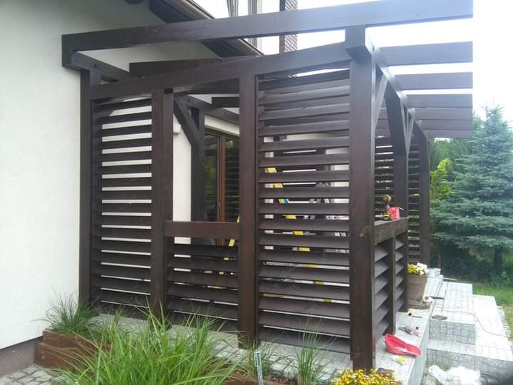 Drewniana pergola z lamelami w kolorze ciemnego brązu, przylegająca do białego budynku, z widocznymi narzędziami i doniczkami na schodach.