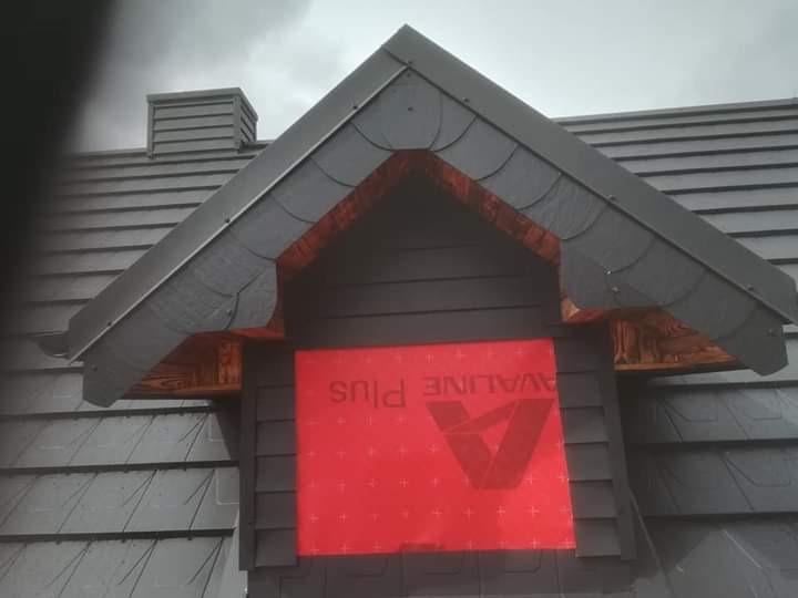 Fragment dachu krytego szarym gontem bitumicznym, widoczny okap z drewnianymi zdobieniami i czerwona folia ochronna z logotypem na ścianie szczytowej, komin w tle.